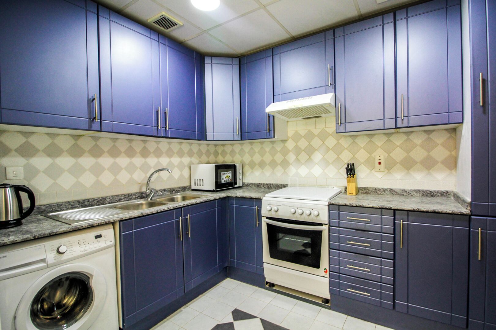 Een kleine keuken met blauwe kastjes, een magnetron, een fornuis, een spoelbak en een wasmachine.
