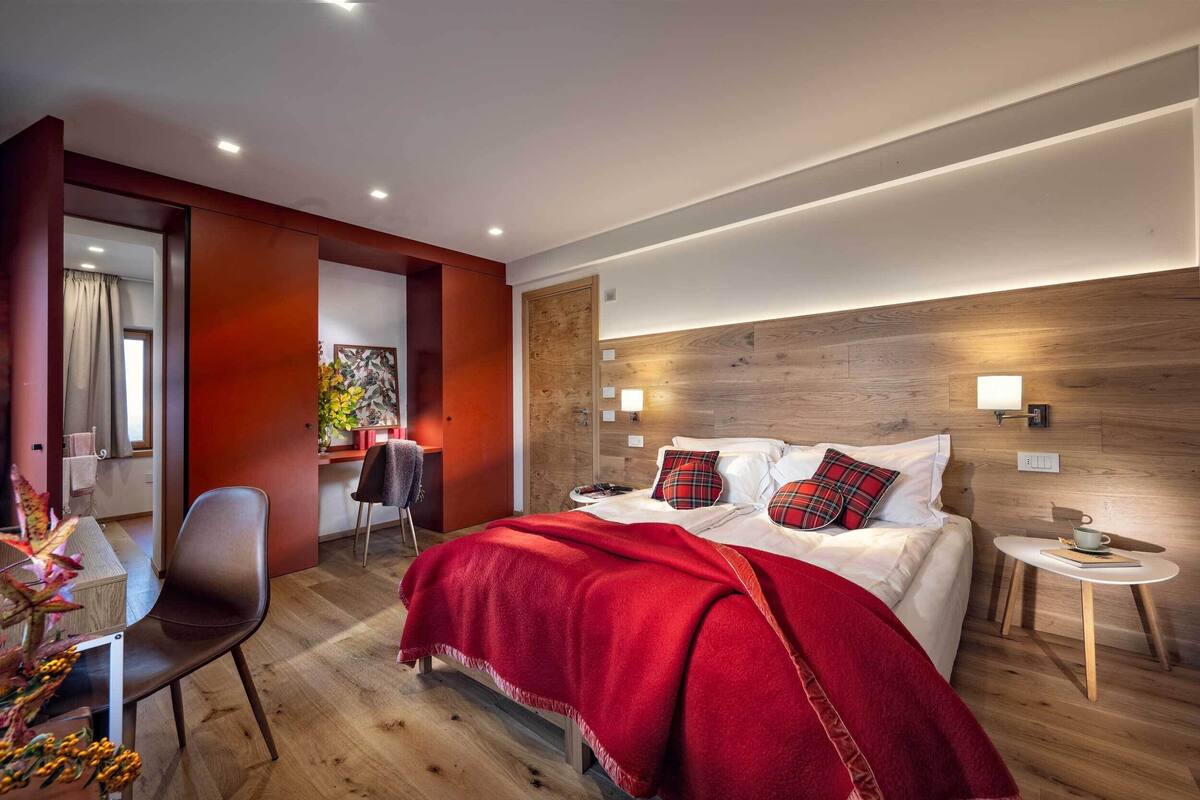 A modern hotel room with a wooden headboard, a red blanket, and a small bedside table with a cup and book.