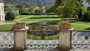 A landscaped garden with a fountain, bordered by a wrought iron fence, and a view of a hill with trees and a building in the background. - Agriturismo Villa di Modolo (Belluno)