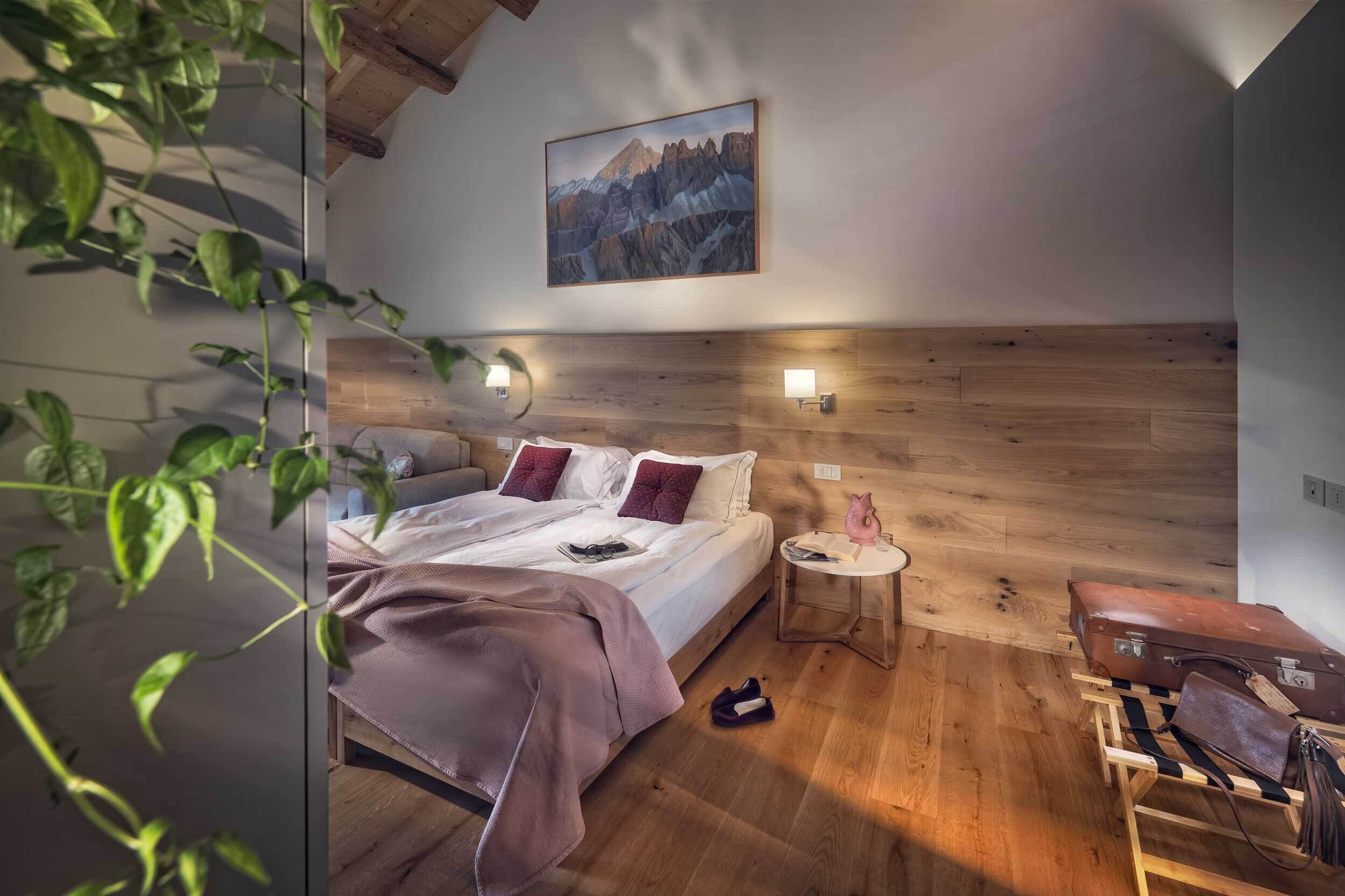 Un dormitorio con cabecera de madera, una cama con manta rosa, dos almohadas, una mesita de noche con lámpara y una maleta sobre un carrito de madera.