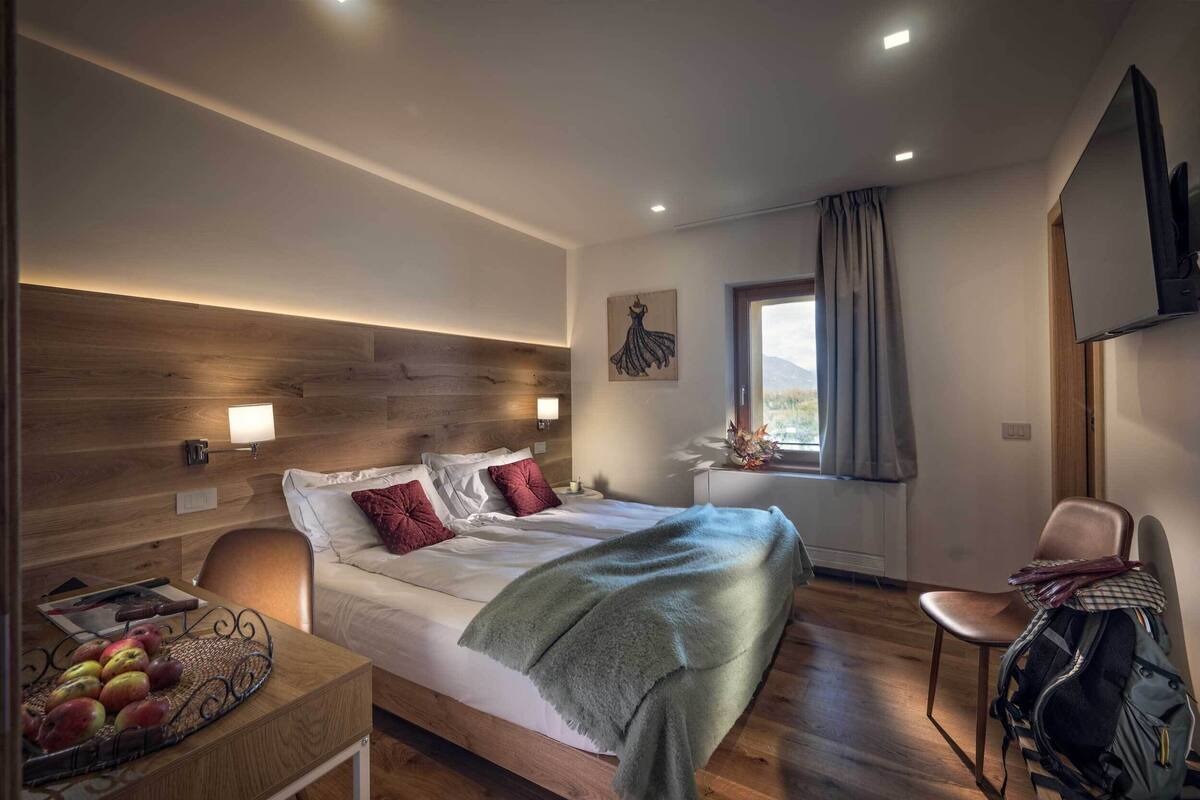 A modern hotel room with a wooden headboard, a bed with white linens, a bedside table with apples, and a chair with a backpack.