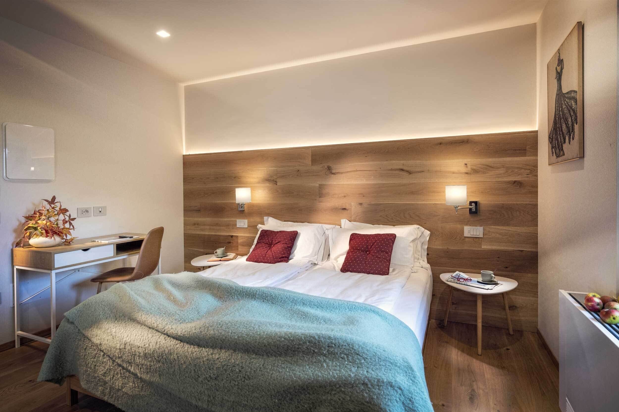 Una habitación de hotel moderna con cabecera de madera, cama con sábanas blancas, un escritorio con silla y una mesita de noche con una taza y una revista.