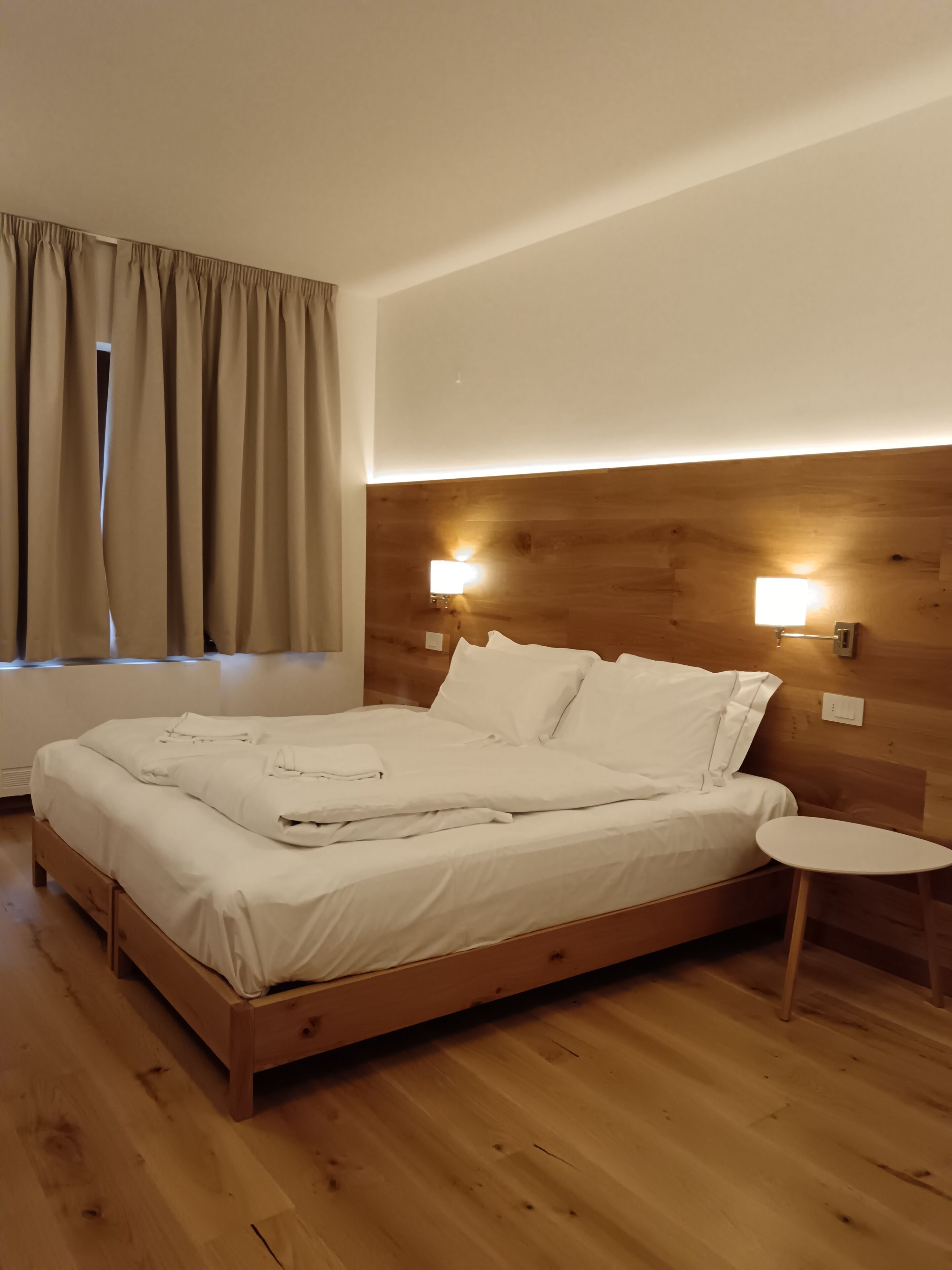 A neatly made bed in a room with wooden flooring, a small bedside table, and two wall-mounted lights.