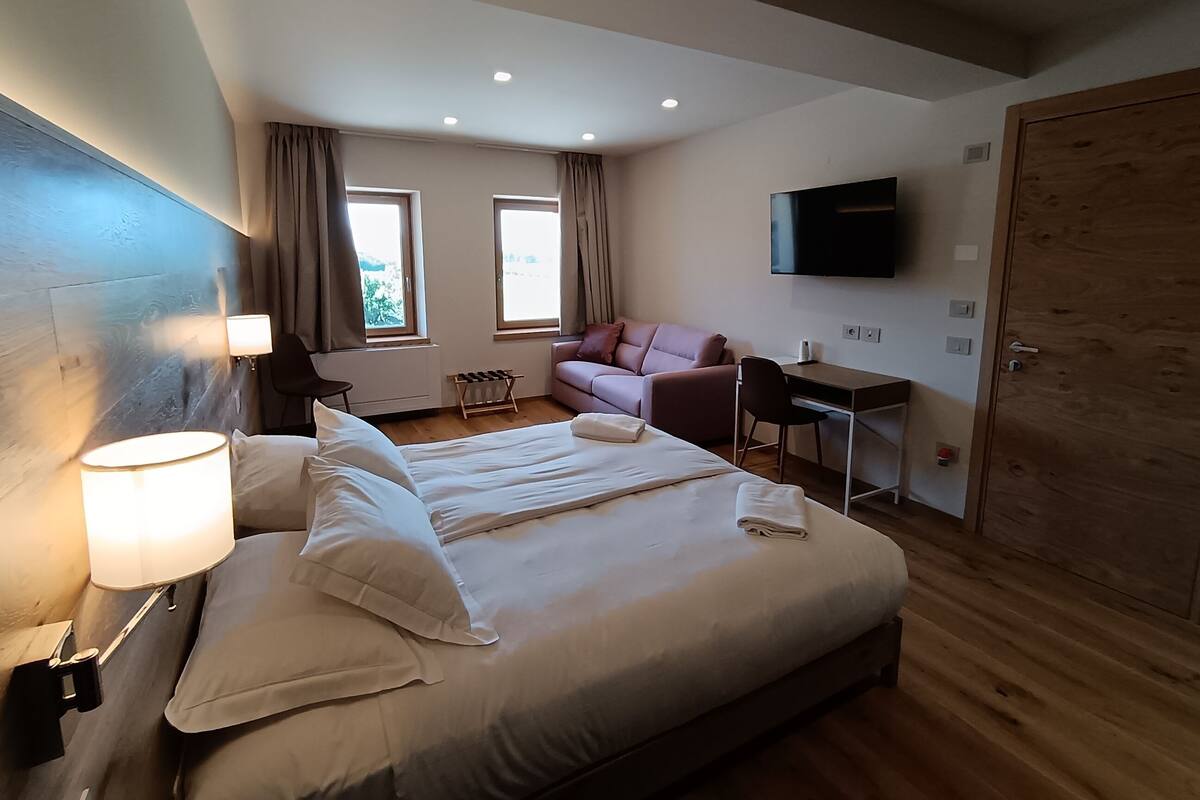 A hotel room with a large bed, a desk, a chair, a TV, and a wardrobe.