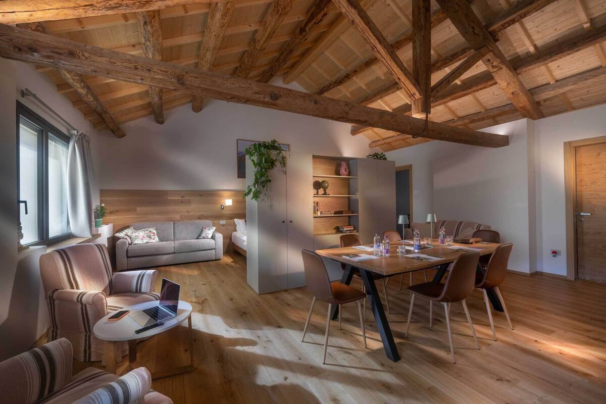 A spacious living area with a wooden ceiling, a dining table with chairs, a sofa, and a small seating area with a laptop.
