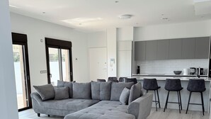 A modern living room with a grey sectional sofa, a white rug, and a kitchen area with grey cabinets and black bar stools.