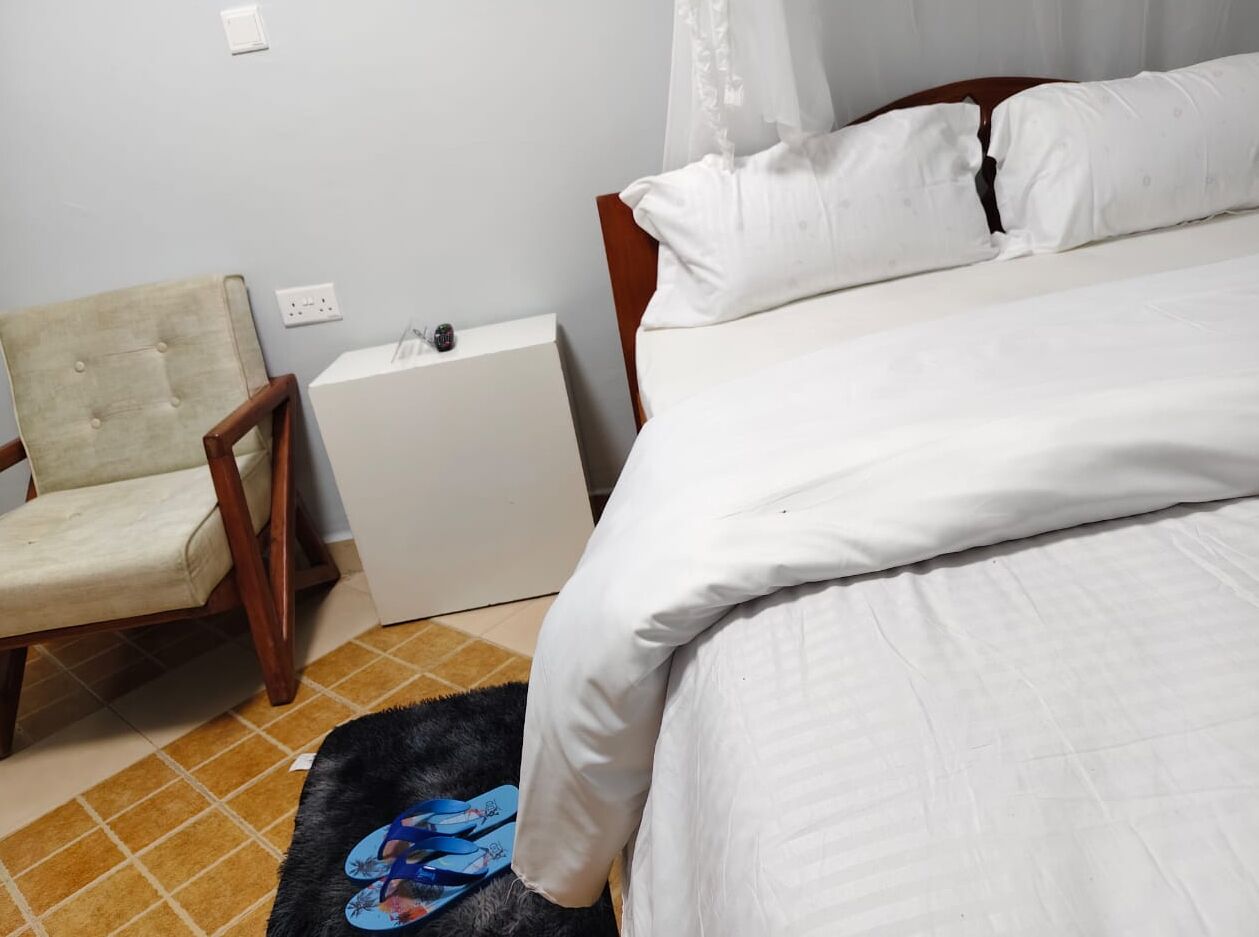 A bedroom with a bed, a chair, a small bedside table, and a power outlet on the wall.