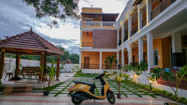 Um pátio com um gazebo, uma scooter e um edifício de vários andares com varandas e um jardim.