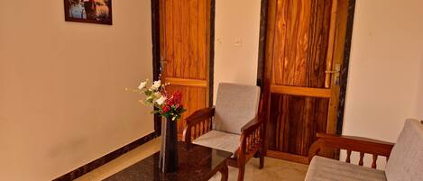 A room with a marble-tiled floor, a wooden table with a vase of flowers, two wooden doors, and a grey sofa.