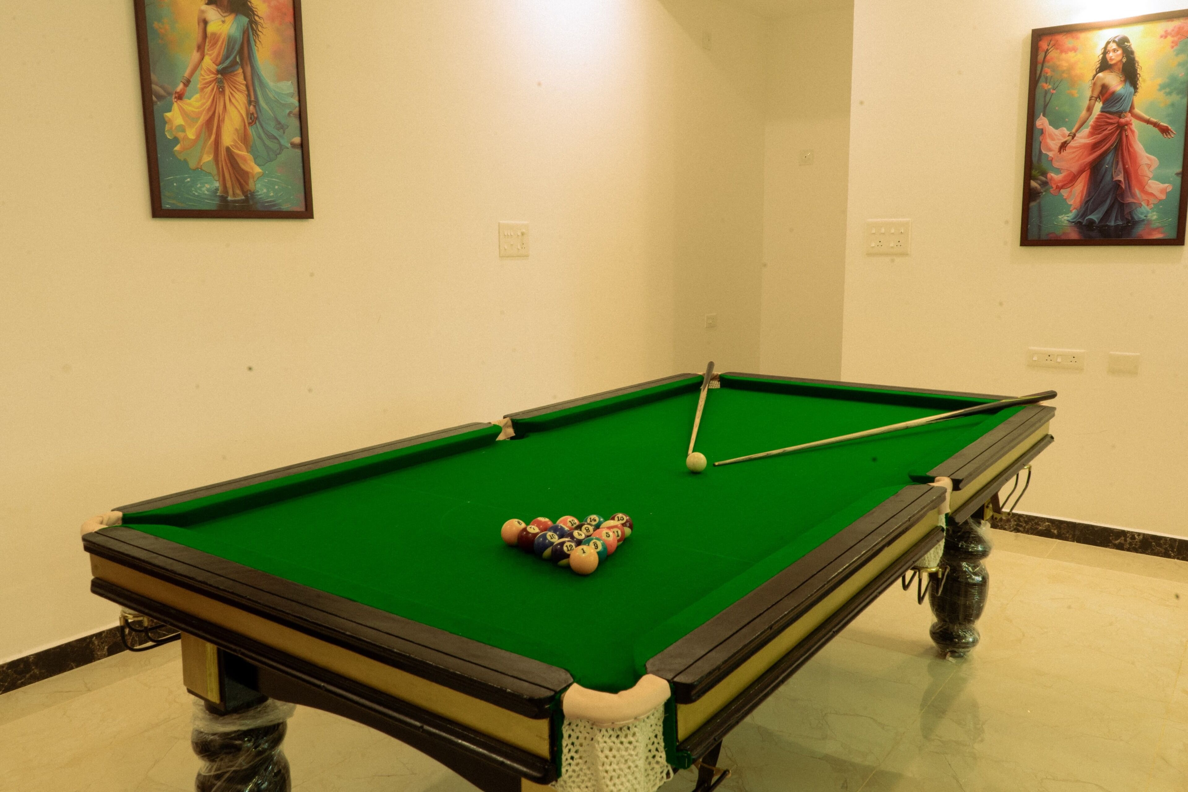 Une table de billard avec des billes, une queue et deux tableaux accrochés aux murs.