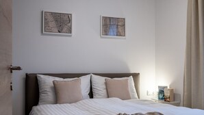 Una cama bien hecha con una colcha a rayas y dos almohadas, una mesita de noche con un libro, y dos mapas enmarcados en la pared.
