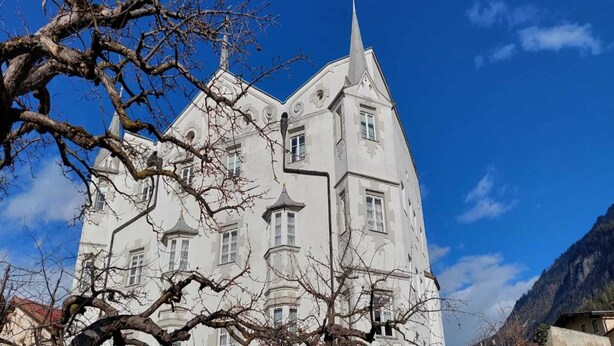 Un edificio grande y blanco de varios pisos, con una torre destacada y un tejado pronunciado.