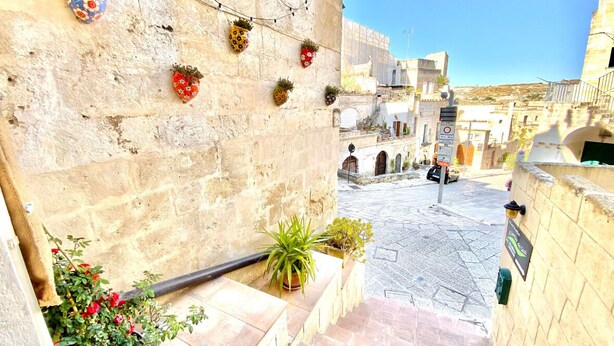 A narrow alleyway with stone walls, potted plants, and a tiled floor leading to a building with a sign. - The Smile of the Sassi Rooms. Idris Superior Deluxe on Sassi view. (Matera)