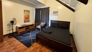 Ein kleiner, gut beleuchteter Raum mit einem Bett, einem Schreibtisch, einem Stuhl, einem Standventilator und einer Wand-Klimaanlage.
