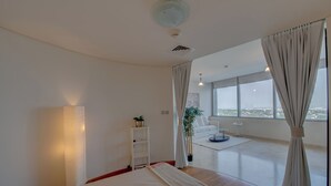A modern hotel room with a large bed, a bedside lamp, a view of the city through large windows, and a potted plant. - Newly launched studio (Dubai)