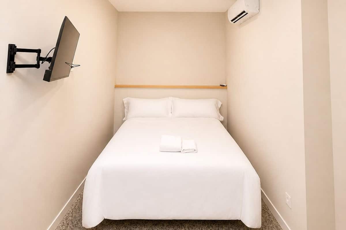 A hotel room with a bed, a mounted TV, an air conditioning unit, and a skylight.