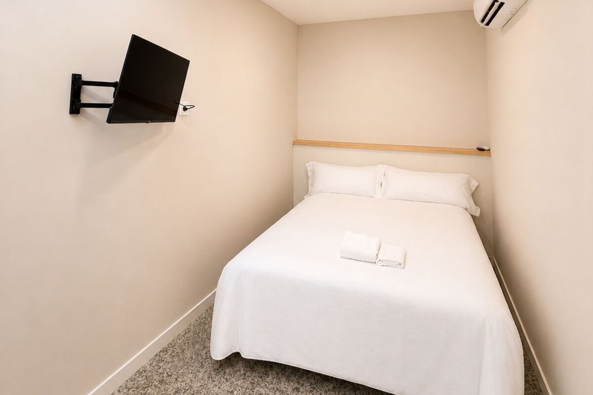 A small, modern hotel room with a bed, a mounted TV, a ceiling light, and a wall-mounted air conditioner.