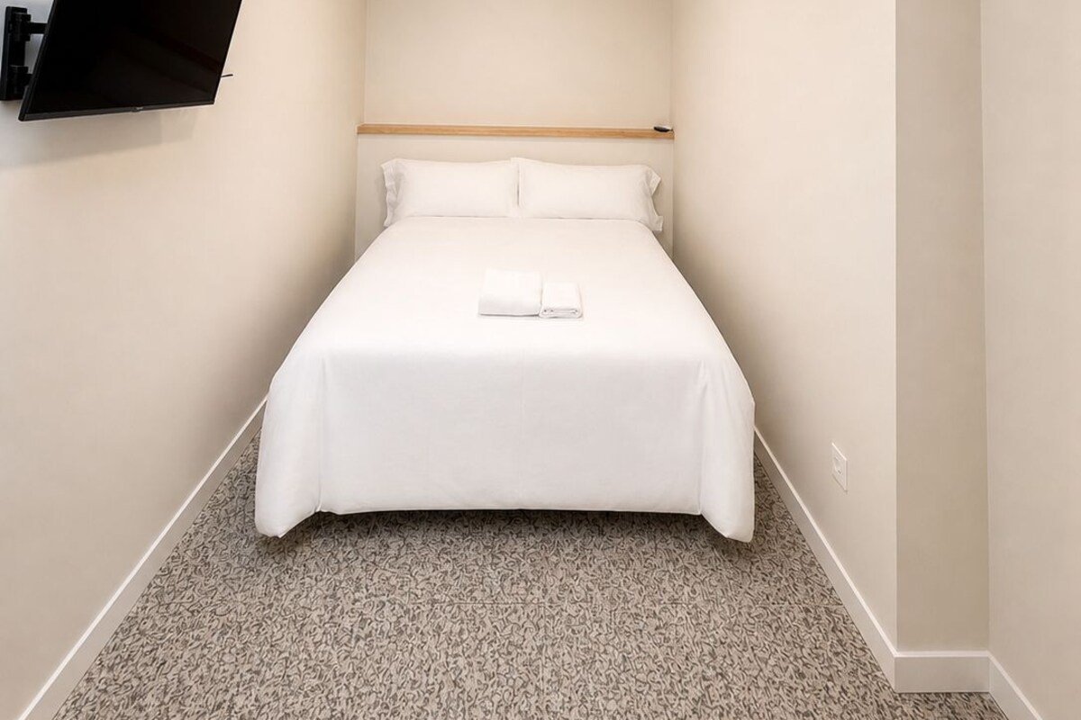 A hotel room with a bed, a mounted TV, an air conditioning unit, and a ceiling light.