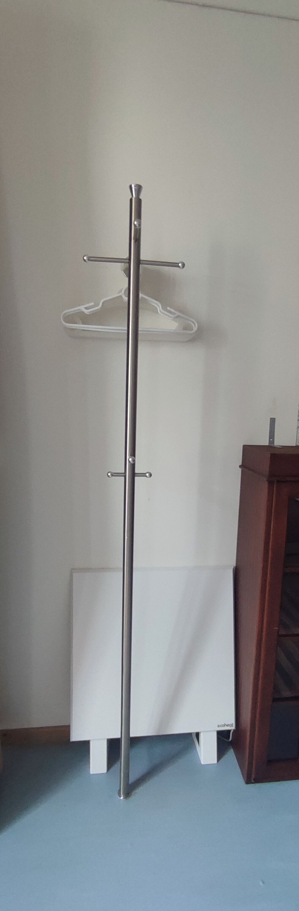 Un grand présentoir métallique à la base blanche, qui ressemble probablement à un porte-manteau, se trouve dans une pièce dont le mur est uni et qui contient une armoire en bois.