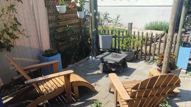 Un patio con sillas y una mesa de madera, rodeado de plantas en macetas y una valla de madera.