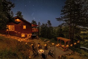 A rustic cabin with a fire pit and outdoor seating, surrounded by trees and a starry night sky. - New 4BR Luxe Getaway Stunning Views/ Hot Tub/ Home Theater & Games- Pet Friendly (Bailey)