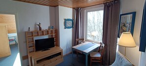 Une pièce avec un téléviseur posé sur un meuble en bois, une table avec des chaises, une fenêtre avec des rideaux et une lampe.