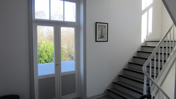 Eine Treppe mit weißen Geländern führt zu einer Zwischenebene, eine Tür mit Glasfeldern und ein gerahmtes Bild an der Wand.