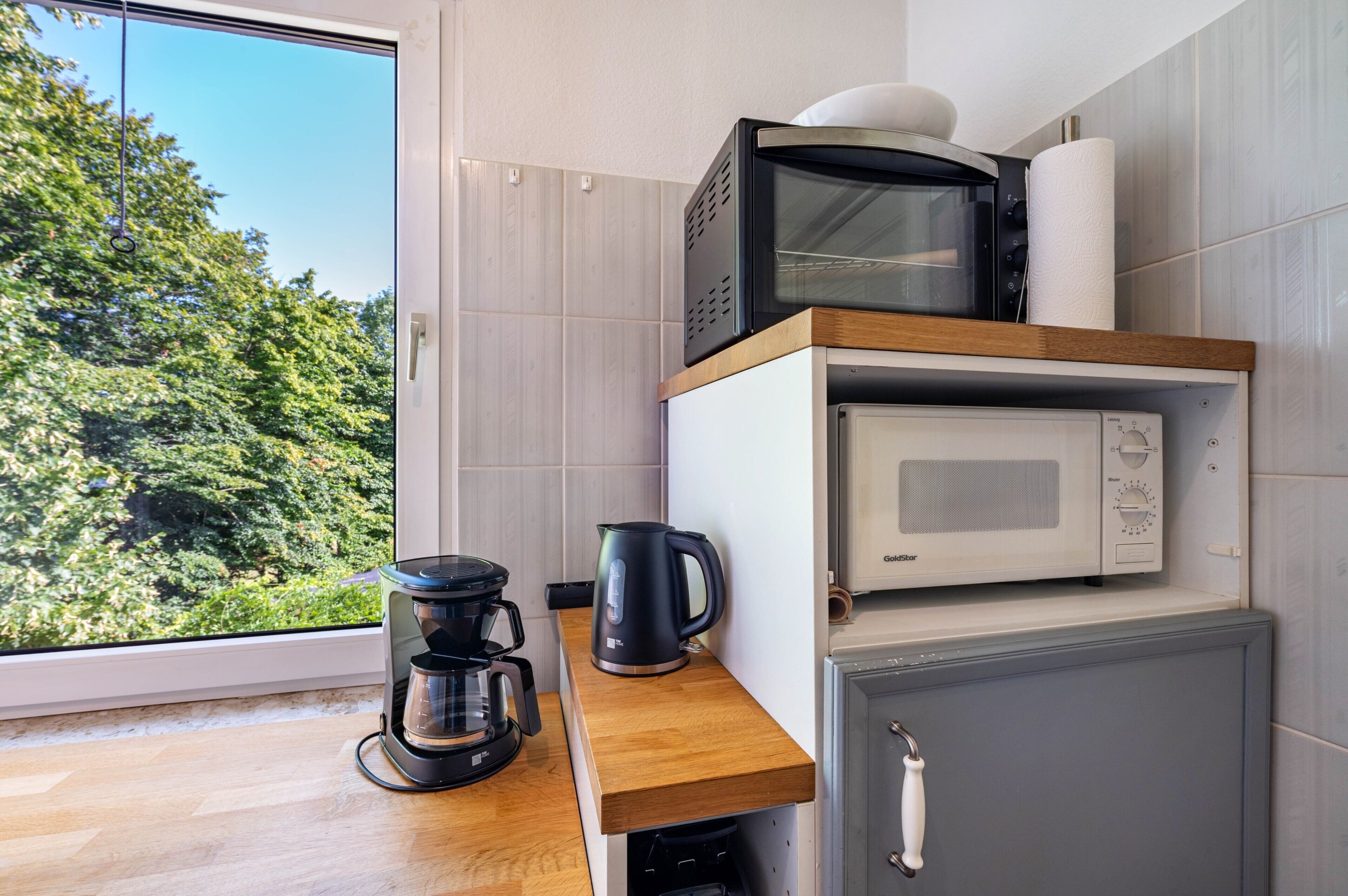 Eine kleine Küche mit Kaffeemaschine, Mikrowelle und Wasserkocher auf einer Holzausziehtischplatte.