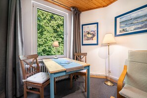 Ein Zimmer mit Tisch und Stühlen, einem Fenster mit Vorhängen, einem Heizkörper, einer Lampe und zwei gerahmten Kunstwerken an der Wand.