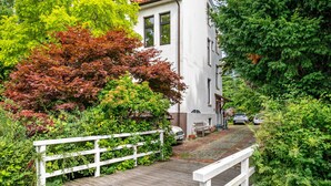 A wooden pathway leading to a white building with a tiled roof, surrounded by lush greenery and a white fence. - Apartment '3 Dg' with Shared Garden and Wi-Fi (Cuxhaven)