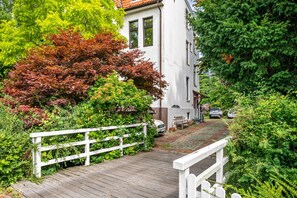 Ein Holzweg führt zu einem weißen Gebäude mit einem Ziegeldach, umgeben von üppiger Vegetation und einem weißen Zaun.