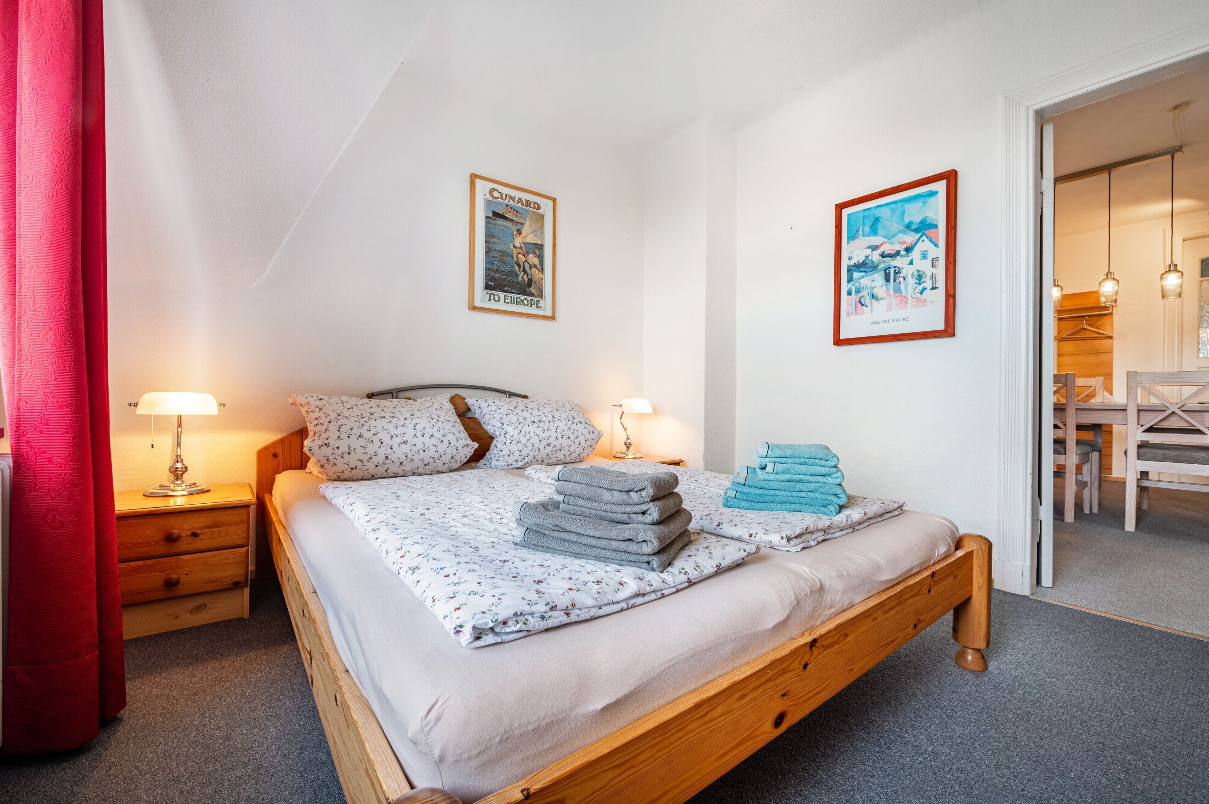 Une chambre à coucher avec un lit en bois, deux tables de chevet avec des lampes, un rideau rouge et deux tableaux encadrés accrochés au mur.