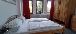 A bedroom with a wooden bed, a radiator, a window with a view of greenery, and a wooden wardrobe.