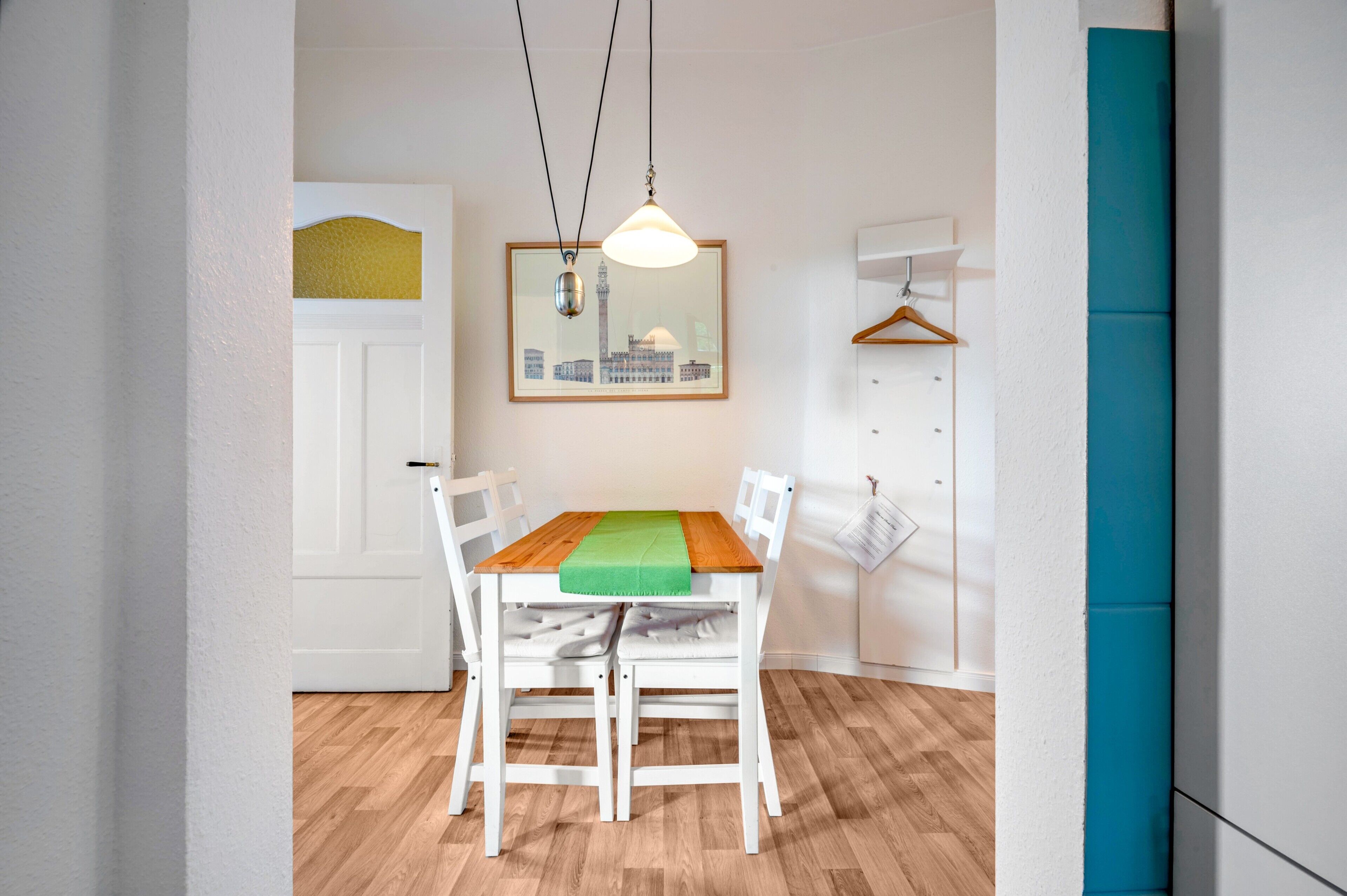 Ein Essbereich mit einem Holztisch, weißen Stühlen, einer hängenden Leuchte und einem gerahmten Stadtbild an der Wand.