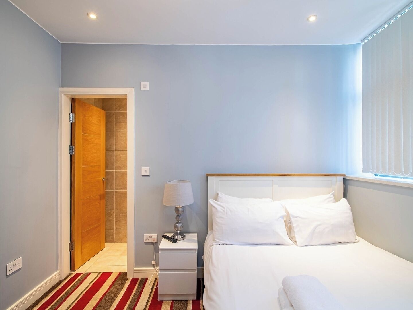 Quarto de hotel com uma cama, mesa de cabeceira, abajur e porta para um banheiro.