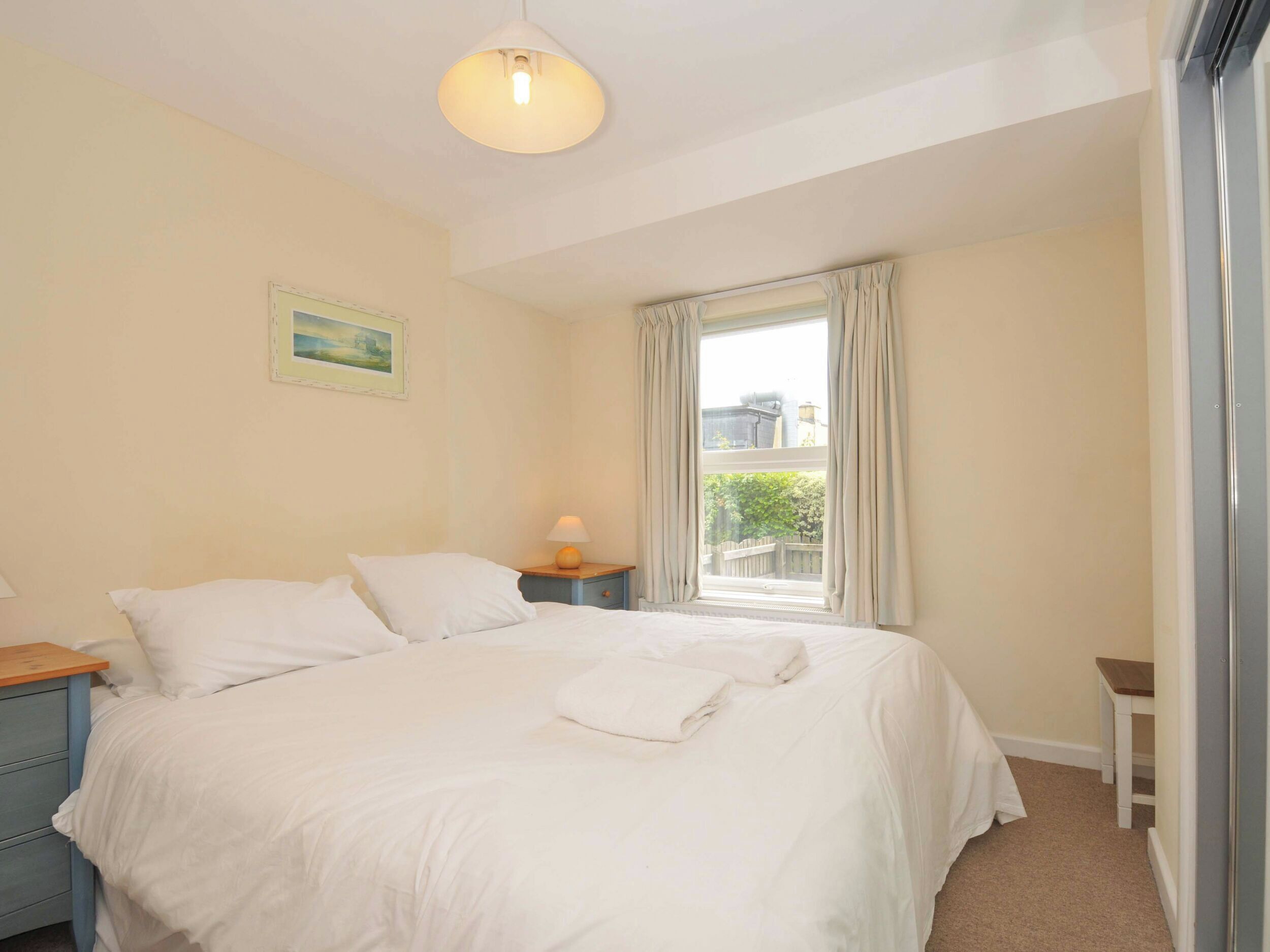 A bedroom with a bed, a nightstand, a lamp, a window with curtains, and a painting on the wall.