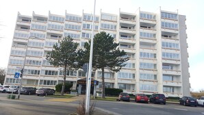 Un edificio de varios pisos con fachada uniforme, un paso de peatones y coches aparcados.