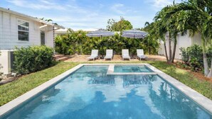 A swimming pool with two lounge chairs and umbrellas, surrounded by palm trees and a white building.