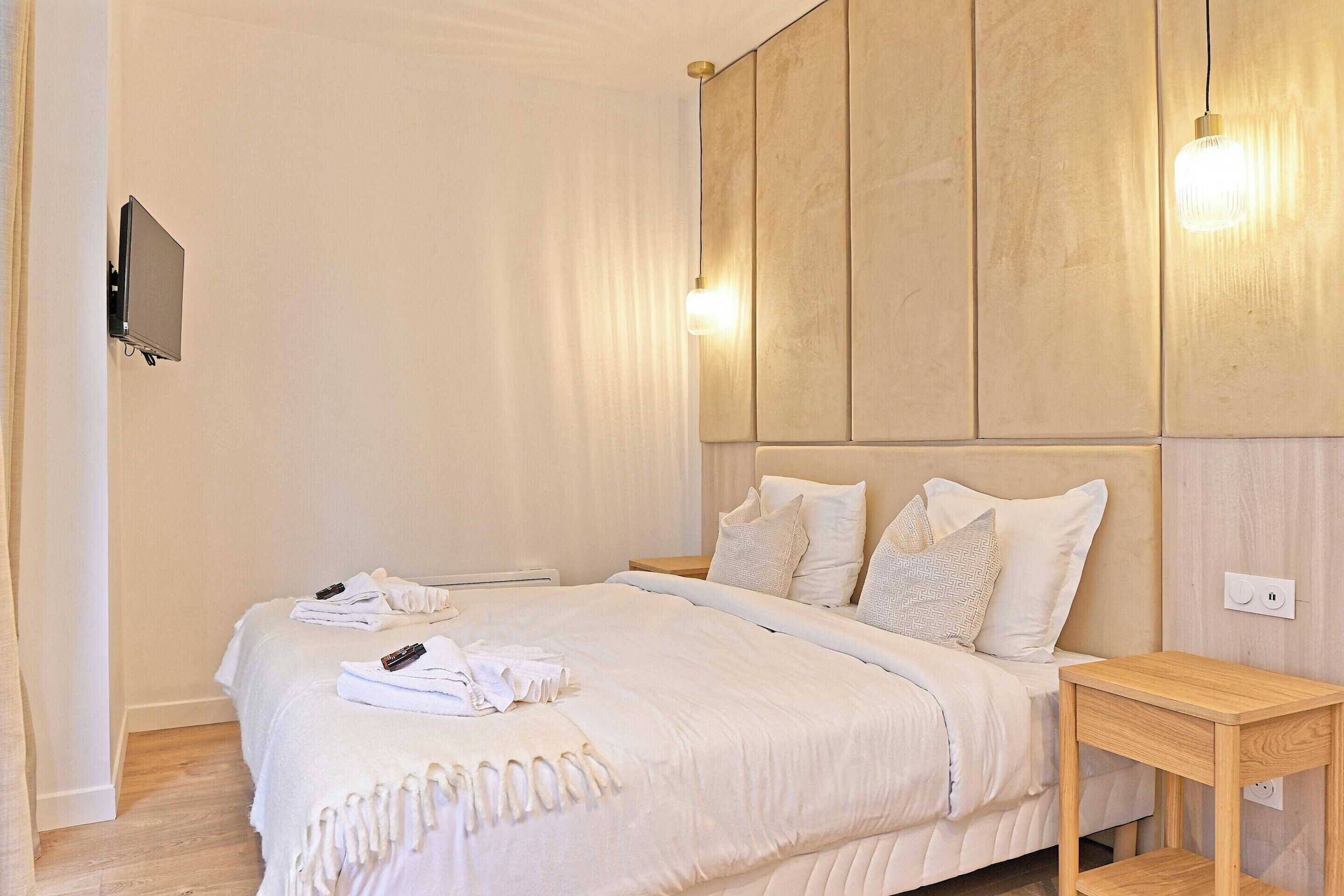 A neatly made bed with white linens and a remote control, a wooden nightstand, and a wall-mounted TV.