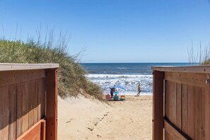 A wooden beach entrance with a sandy path leading to the sea, a beach umbrella, and beach chairs.
