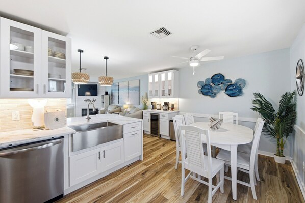A modern kitchen with white cabinets, a stainless steel dishwasher, a built-in oven, and a dining area with white chairs and a table.