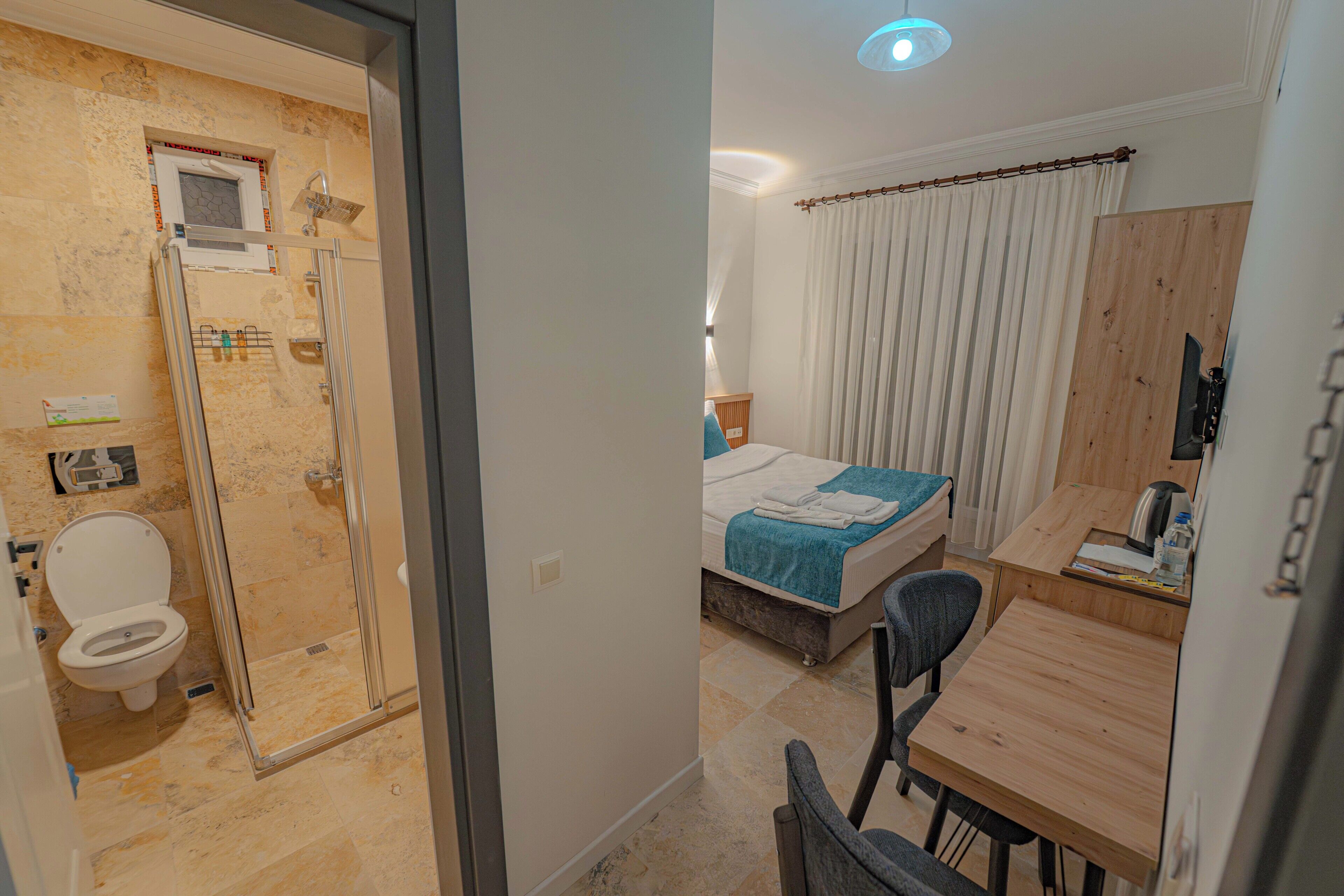 A hotel room with a shower, toilet, bed, bedside table, chair, and a desk with a chair.