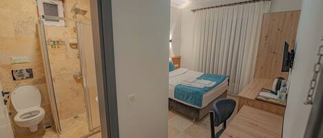 A hotel room with a shower, toilet, bed, bedside table, chair, and a desk with a chair.