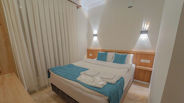 A hotel room with a bed, wooden headboard, two wall-mounted lights, a window with blinds, and a wooden table. - Full Of Life (Nevşehir)