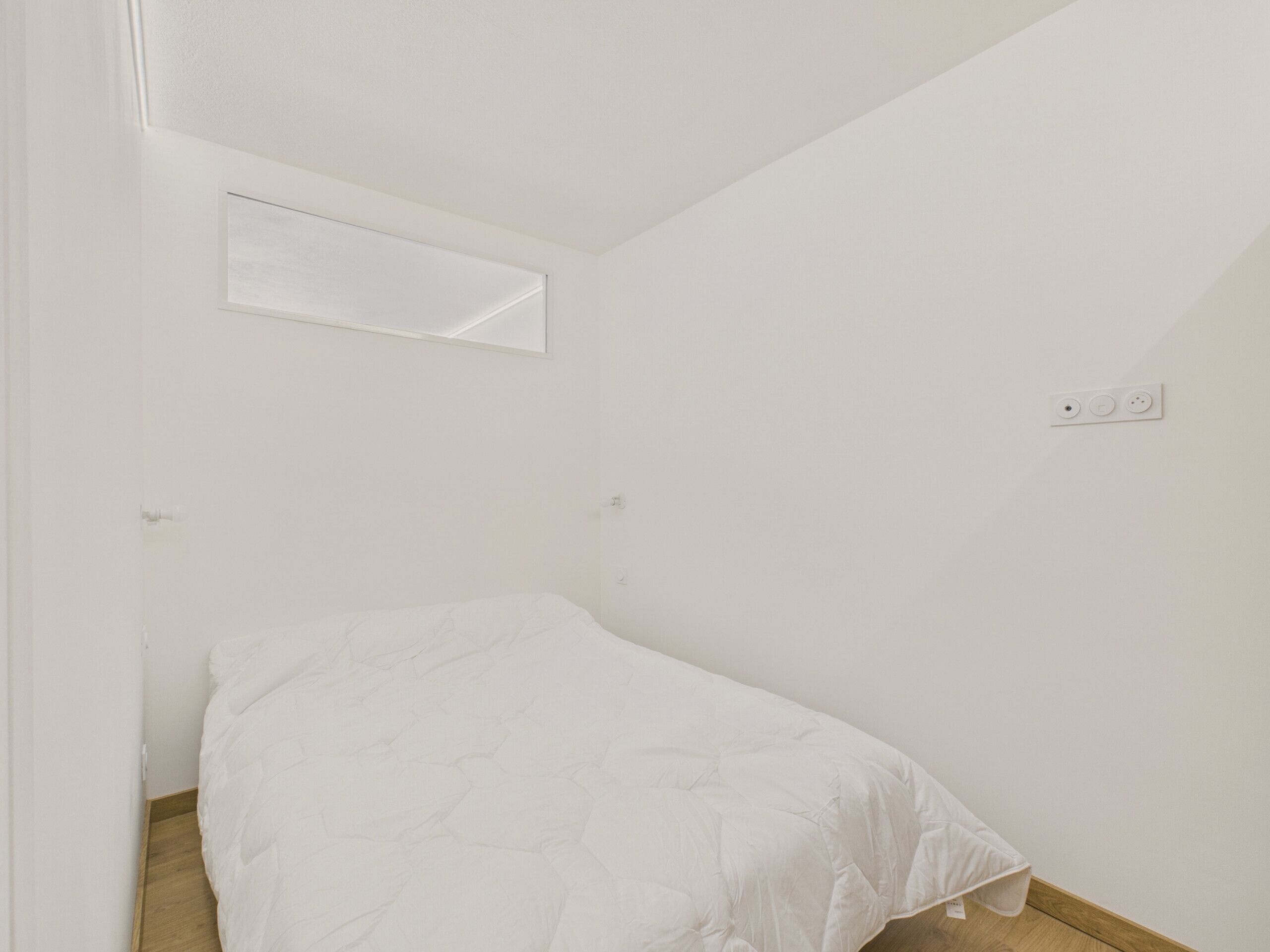 Una habitación pequeña con cama individual, piso de madera, pared blanca y una ventana pequeña.