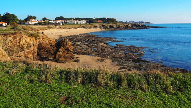 Un paisaje costero con una playa arenosa, un litoral rocoso y casas al fondo.