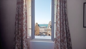 Une vue à travers une fenêtre ornée de rideaux à motifs, dévoilant une ville côtière avec des bâtiments historiques et un ciel dégagé.