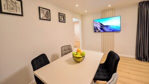 Un coin repas avec une table, des chaises et un panier de fruits. Des tableaux encadrés ornent le mur et un téléviseur est fixé au-dessus de la table.