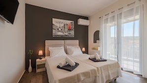 A hotel room with two beds, a painting on the wall, an air conditioning unit, a nightstand with a lamp, and a view of the outside through large windows. - 3a's Family Luxury Villa (Rethymno)