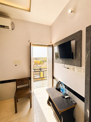 Habitación de hotel con una televisión montada en la pared, una mesita con una tetera y botellas, un banco de madera y una puerta que da a un balcón con vistas a edificios.